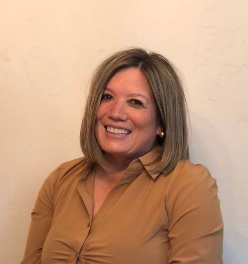 The image features a woman sitting in front of a white wall. She has short, light-colored hair and is wearing a dark blazer over a lighter top. Her expression is neutral with a slight smile.