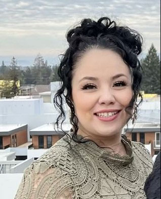 The image features a woman with dark hair styled in waves, wearing makeup and a sleeveless top with lace detailing. She is smiling at the camera and appears to be outdoors during daylight hours.