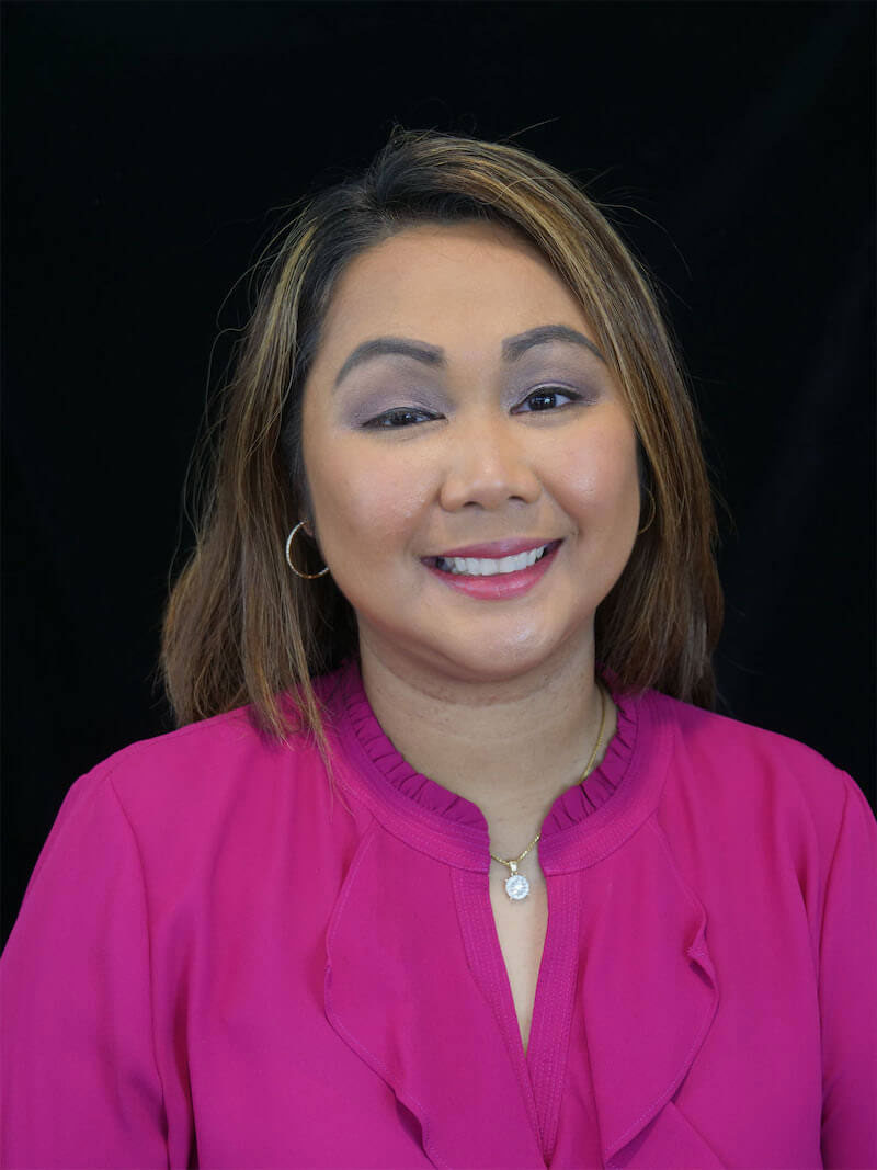 The image shows a smiling woman with brown hair, wearing a pink top and sitting against a dark background.