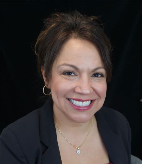 The image shows a woman with medium-length hair, wearing a gold necklace and a black top. She has a smile on her face and is looking directly at the camera.
