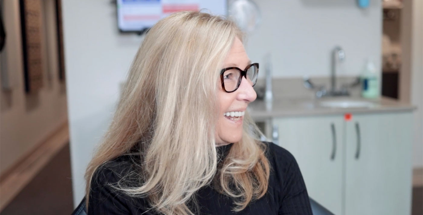 The image shows a woman with short blonde hair and glasses, smiling at the camera. She appears to be in an indoor setting, possibly a dental or medical office, as suggested by the background that includes a sink and countertop.