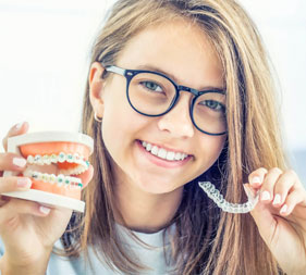 The image shows a young woman with glasses holding up a dental model and a toothbrush, smiling at the camera.