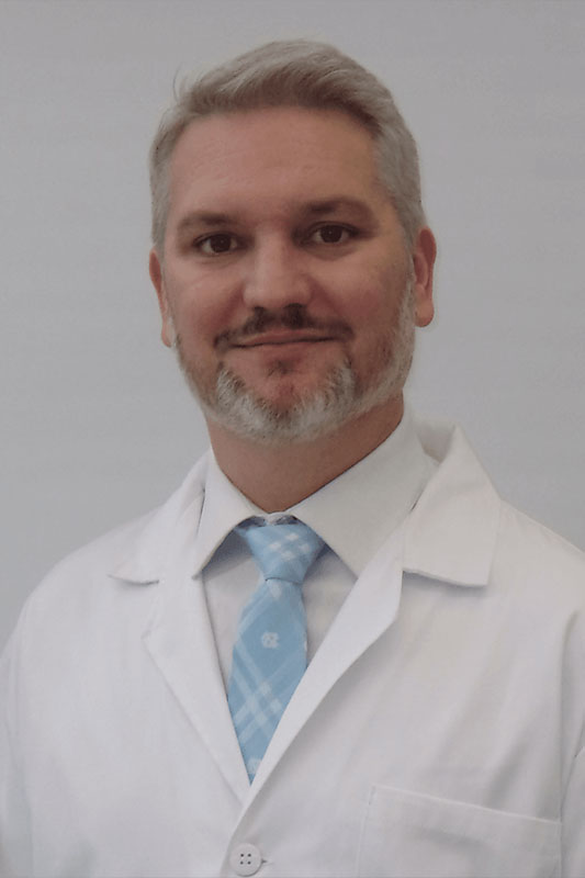 The image shows a man with facial hair, wearing a white lab coat and a blue tie, standing in front of a plain background. He appears to be a professional, possibly a scientist or researcher, given his attire and the setting.