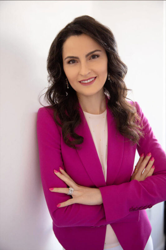 A woman in a pink blazer stands confidently with her hands on her hips, wearing a white top and gold earrings.