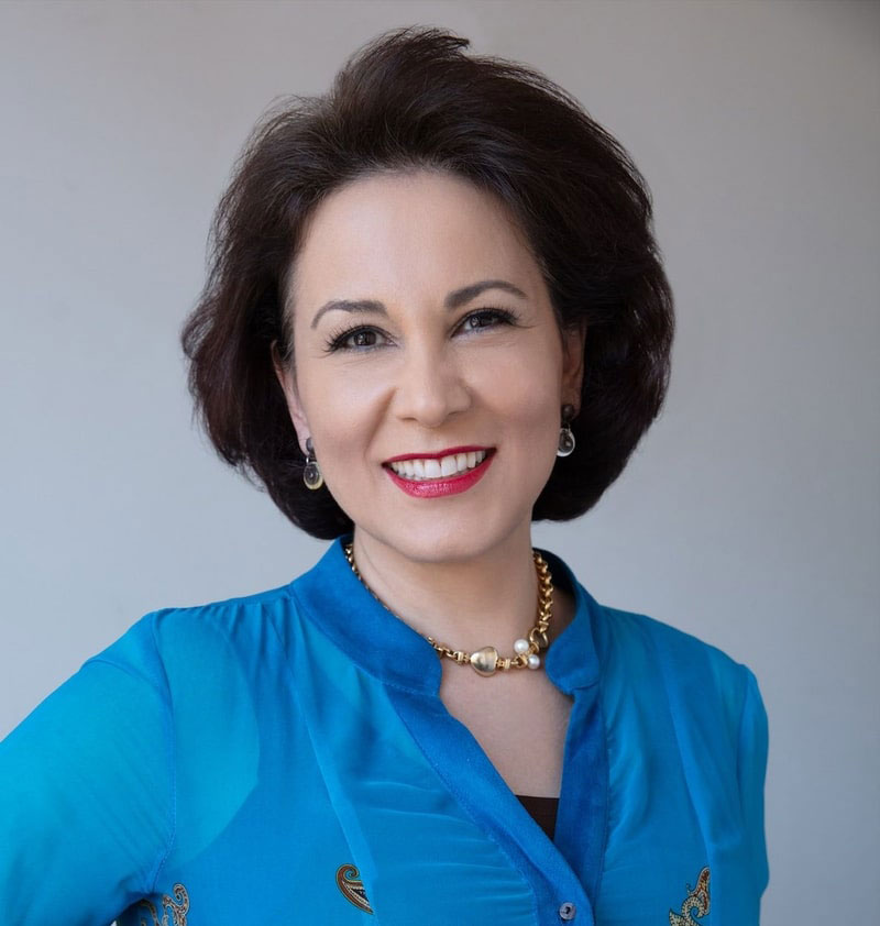 The image is a portrait of a woman posing for the camera. She has dark hair, wears a blue top and a necklace, and is smiling while looking directly at the camera.