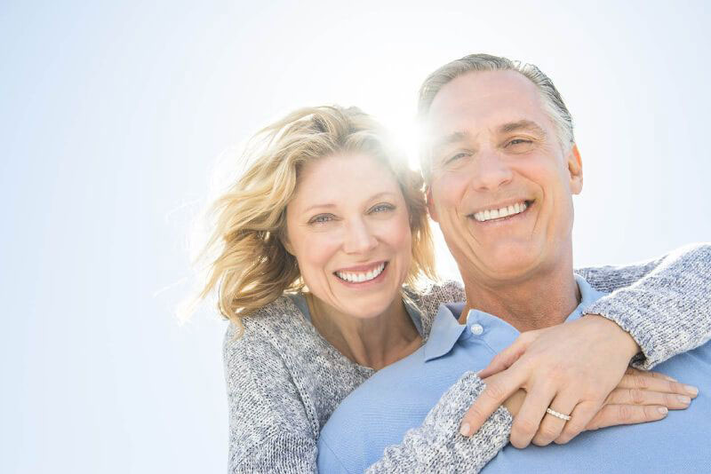 An elderly couple embracing and smiling at the camera, with a bright sunny background.