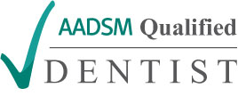 The image is a logo or certification mark with text. It features a checkmark symbolizing approval, and the text includes  AADSM Qualified Dentist,  indicating that a dentist has met certain qualifications set by the American Academy of Dental Sleep Medicine.