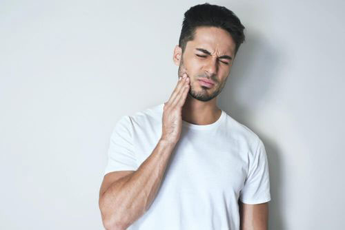 A young man with a beard and mustache, wearing a white t-shirt, is captured in a close-up shot against a plain background. He appears to be looking upwards with his mouth slightly open, possibly in the midst of speaking or singing.