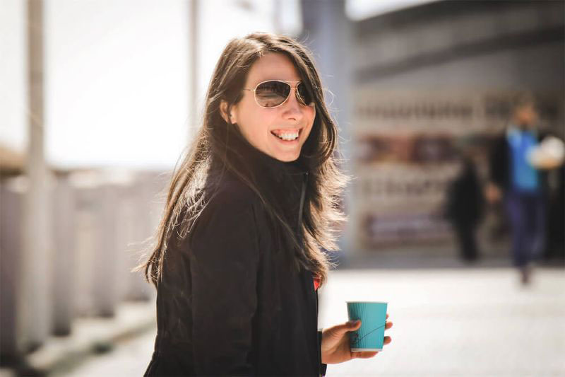 The image is a photograph of a woman standing outdoors, smiling slightly, holding a cup in her left hand. She has sunglasses on top of her head and is wearing a dark jacket with a light undershirt. The background shows an urban setting with a building and other people visible in the distance.