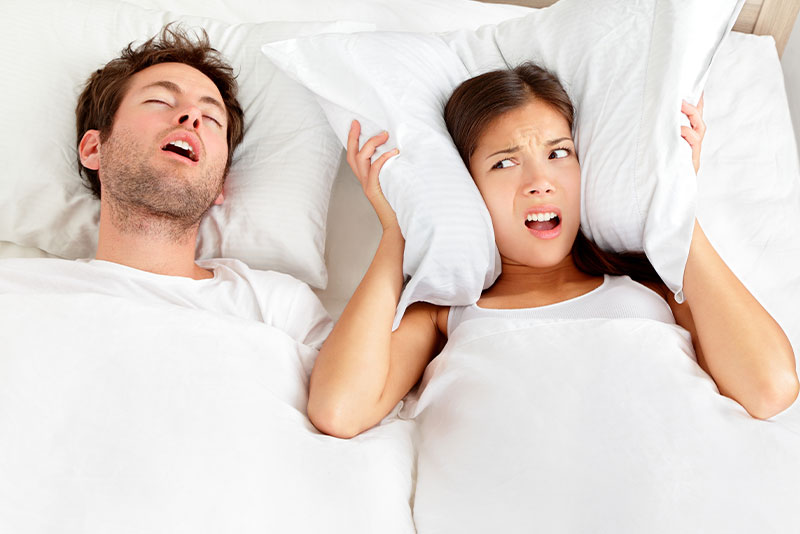 Two people in bed, one with a pillow over their head and the other looking surprised or concerned.