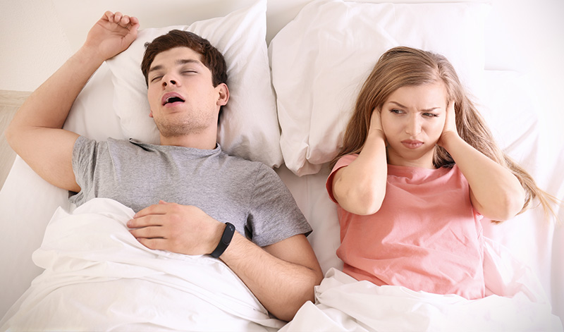 A young couple sleeping in bed, with the man on his back and the woman resting her hand on her ear.