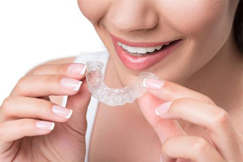 A woman holding a clear plastic dental retainer to her mouth, smiling and showcasing the product.