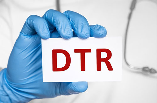 The image features a pair of gloved hands holding up a white card with the letters  DTR  in red, against a blurred background that appears to be a medical setting.