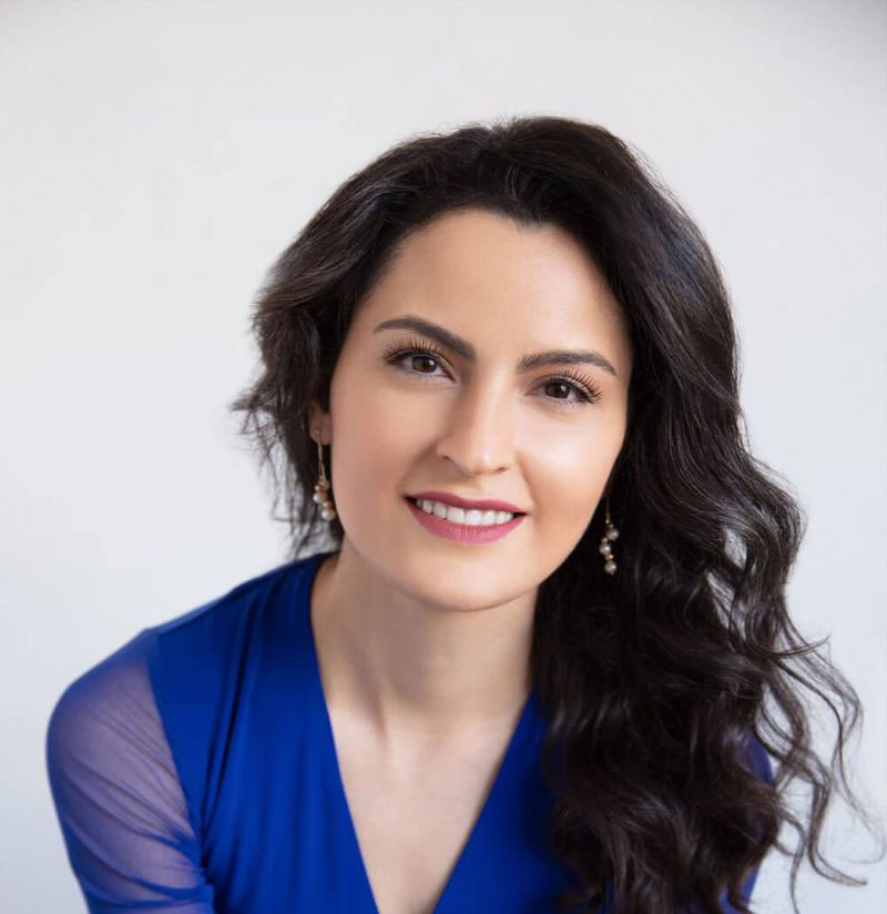 A woman with long dark hair posing for a portrait, wearing a blue top and earrings.