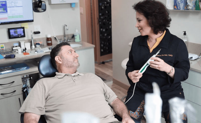 The image shows a dental office setting with two individuals; one is seated in the dentist's chair while another stands by, possibly a dental professional.