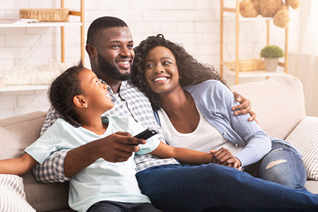 This is a color photograph featuring a family of four, including an adult male, an adult female, and two young children. They appear to be in a casual, indoor setting with a couch and decorative elements in the background. The man is smiling and looking at the camera, while the woman has her arm around him and is also smiling.