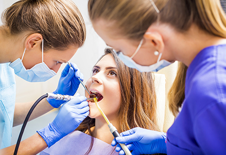 The image depicts a dental hygiene scene with a patient receiving treatment from a dentist, assisted by a dental hygienist, all wearing protective gear.