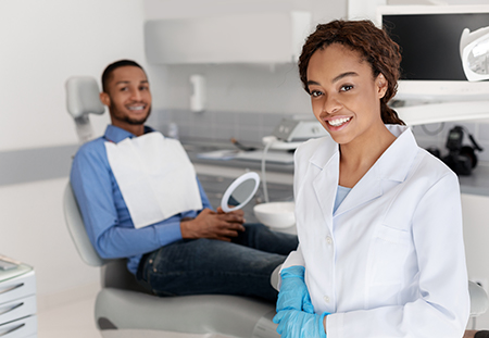 A dental hygienist in a white coat and blue gloves is smiling at the camera, standing behind a dental chair where an adult patient sits, with another person in the background.