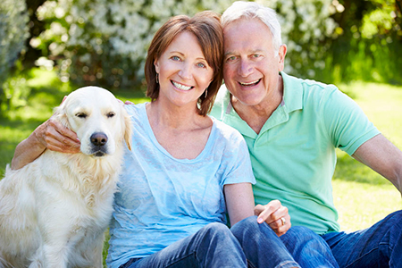 An elderly couple sitting on the grass with a golden retriever, smiling and looking at the camera.