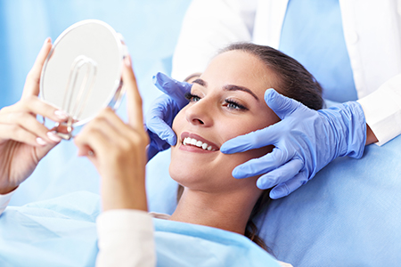 A woman is lying on a dental chair, holding up a magnifying mirror to inspect her teeth while a dentist in protective gloves examines her mouth.