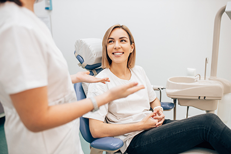 A woman in a dental chair is smiling at another woman, who is standing and gesturing towards the seated woman with her hands.
